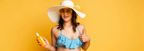 Молодая женщина в солнечных очках и соломенной шляпе держит солнцезащитный крем и показывает большой палец вверх на желтом, горизонтальном изображении — стоковое фото