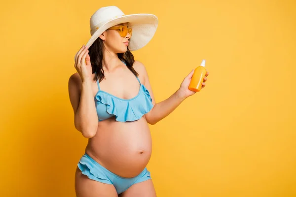Young pregnant woman in swimsuit touching straw hat and holding sunscreen on yellow — Stock Photo