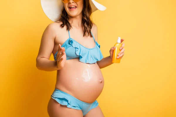 Cropped view of pregnant woman applying sunscreen on tummy while posing on yellow — Stock Photo
