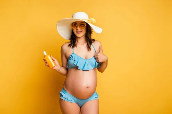 Young pregnant woman in swimsuit holding sunscreen and showing shrug gesture on yellow — Stock Photo