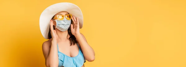 Prise de vue panoramique de la femme en chapeau brut touchant lunettes de soleil et masque médical sur jaune — Photo de stock