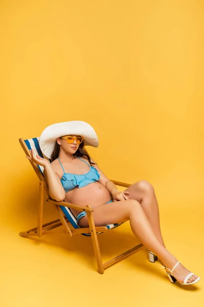Jeune femme enceinte en chapeau de paille et maillot de bain bleu relaxant en transat sur jaune — Photo de stock