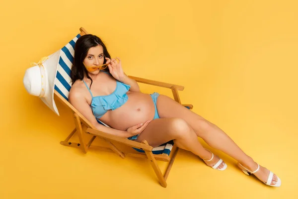Vista ad alto angolo della donna incinta guardando la fotocamera e toccando gli occhiali da sole mentre seduto sulla sedia a sdraio sul giallo — Foto stock