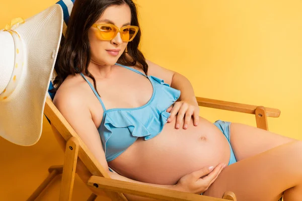 Mulher grávida em óculos de sol olhando para longe e tocando barriga enquanto toma banho de sol em cadeira de praia em amarelo — Fotografia de Stock
