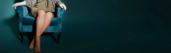 Ausgeschnittene Ansicht einer modischen Schwangeren, die mit überkreuzten Beinen im Veloursessel auf dunkelblauer, panoramischer Aufnahme sitzt — Stockfoto