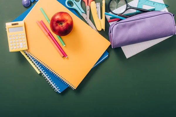 Верхний вид спелых яблок и войлочных ручек на разноцветных копировальных книгах возле школьных канцелярских принадлежностей на зеленой доске — стоковое фото