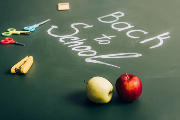 Messa a fuoco selettiva di deliziose mele vicino a iscrizione scolastica e forniture scolastiche sulla lavagna verde — Foto stock