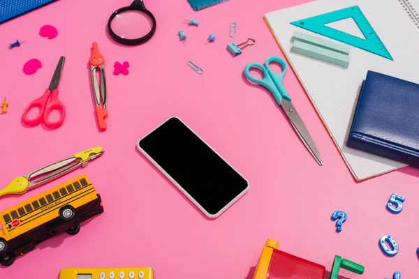 Vista de ángulo alto de teléfono inteligente con pantalla en blanco cerca del modelo de autobús escolar y papelería en rosa - foto de stock