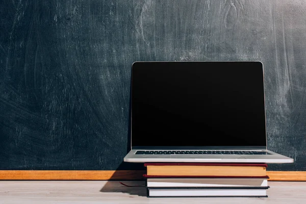 Laptop with with blank screen on stack of textbooks near black chalkboard — Stock Photo