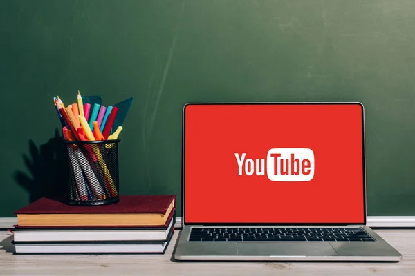 KYIV, UKRAINE - JULY 7, 2020: Laptop with Youtube website near pen holder with stationery on stacked books near green chalkboard — Stock Photo