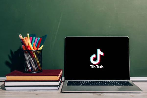 KYIV, UKRAINE - JULY 7, 2020: Laptop with Tik Tok website near pen holder with stationery on stacked books near green chalkboard — Stock Photo