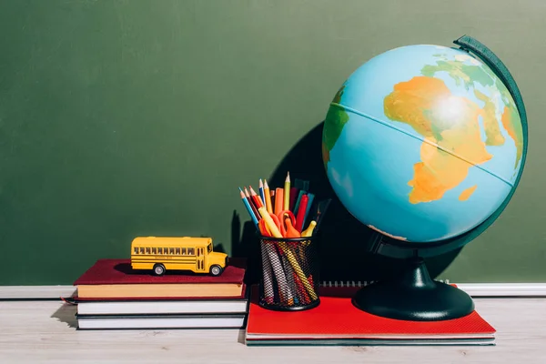 Globus und Stifthalter auf Notizbuch in der Nähe des Schulbusmodells auf Büchern in der Nähe der grünen Tafel — Stockfoto