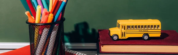 Horizontal image of school bus model on books near pen holder with stationery on notebook near green chalkboard — Stock Photo