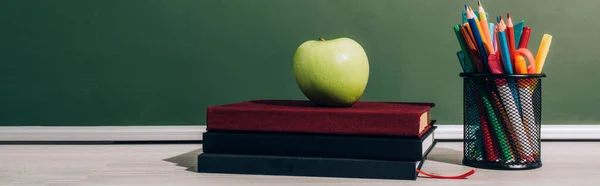 Imagem horizontal de maçã madura em livros perto de porta-caneta com lápis de cor perto de quadro-negro verde — Fotografia de Stock