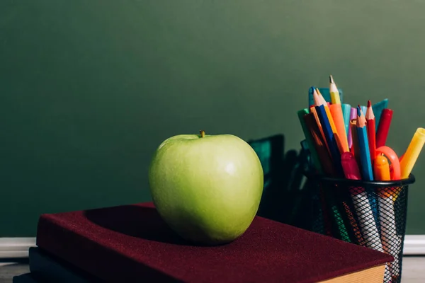 Целое яблоко на книгах возле держателя ручки с цветными карандашами и войлочными ручками на столе рядом с зеленой доской — стоковое фото
