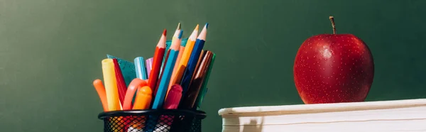 Encabezado del sitio web de titular de la pluma con lápices de color y bolígrafos, y manzana madura en los libros cerca de pizarra verde - foto de stock