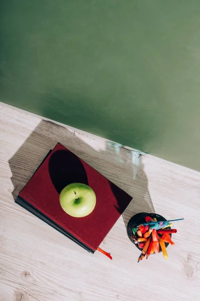 High angle view of whole apple on books near pen holder with stationery on desk near green chalkboard — Stock Photo