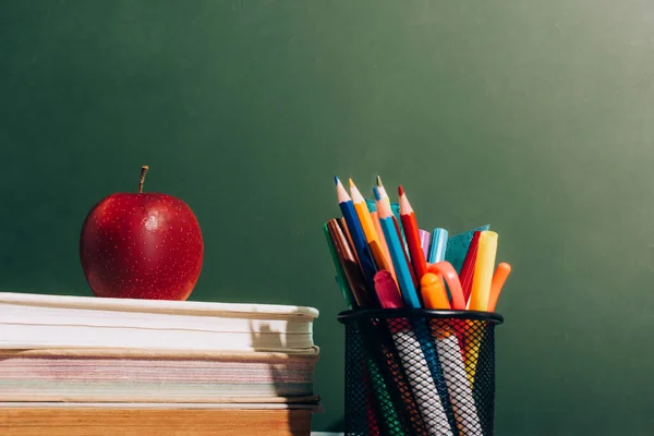 Pen holder with color pencils and felt pens and ripe apple on books near green chalkboard — Stock Photo