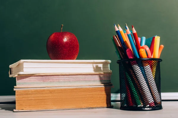 Спелое яблоко на книгах и ручке с цветными карандашами и войлочными ручками на столе рядом с зеленой доской — стоковое фото