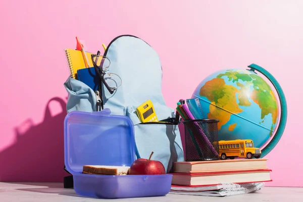 Sac à dos scolaire plein de papeterie près de la boîte à lunch, globe, modèle d'autobus scolaire et livres sur rose — Photo de stock