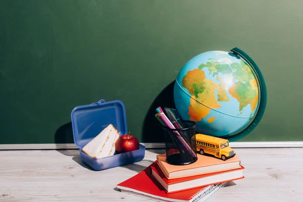 Земной шар возле коробки для обеда, книги, держатель ручки и школьный автобус модель один стол рядом с зеленой доской — стоковое фото