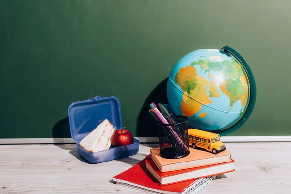 Globe near lunch box, books, pen holder and school bus model one desk near green chalkboard — Stock Photo