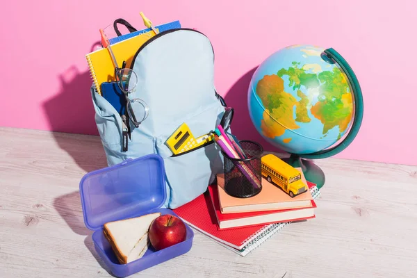 Blauer Rucksack mit Schulbedarf in Erdnähe, Lunchbox, Büchern, Stiftehalter und Schulbusmodell auf rosa — Stockfoto