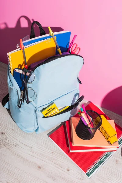 Vue grand angle du sac à dos plein de fournitures scolaires près des livres, porte-stylo et modèle d'autobus scolaire sur rose — Photo de stock