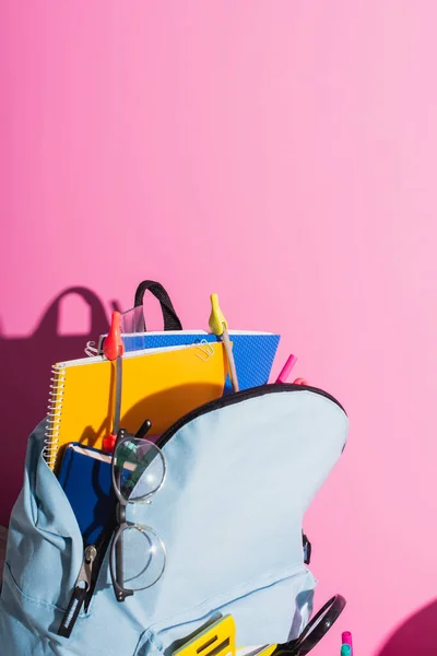 Синий рюкзак с блокнотами, школьными принадлежностями и очками на розовом — стоковое фото