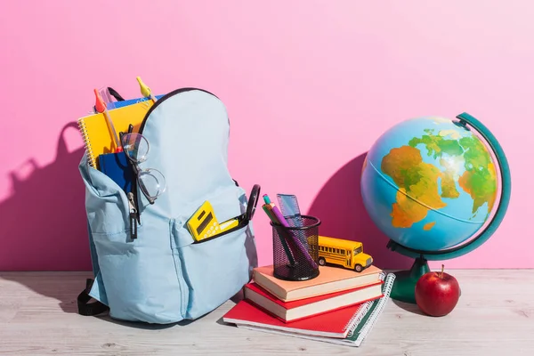 Mochila azul con útiles escolares cerca del mundo, libros, portalápices, manzana fresca y modelo de autobús escolar en rosa - foto de stock