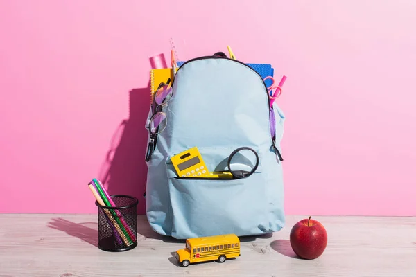 Mochila azul embalada com artigos de papelaria escolar perto do modelo de ônibus escolar, livros, maçã madura e suporte de caneta em rosa — Fotografia de Stock
