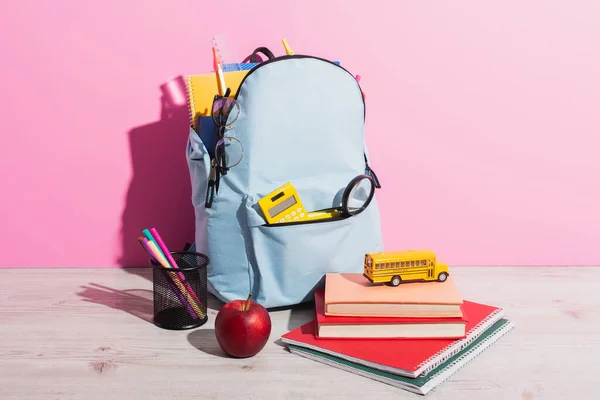 Mochila completa com artigos de papelaria escolar perto de ônibus escolar de brinquedo em livros, maçã madura e suporte de caneta em rosa — Fotografia de Stock