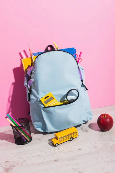 Schulrucksack mit Schreibwaren in der Nähe des Schulbusmodells, ganzer Apfel und Halter mit Filzstiften auf rosa — Stockfoto