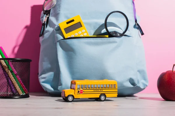 Blue backpack with school supplies near school bus model, ripe apple and pen holder with felt pens on pink — Stock Photo