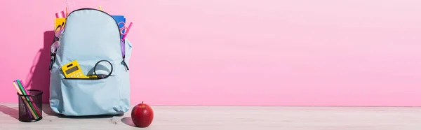 Horizontales Bild eines blauen Rucksacks mit Schulbedarf in der Nähe des ganzen Apfels und Stifthalter mit Filzstiften auf rosa — Stockfoto