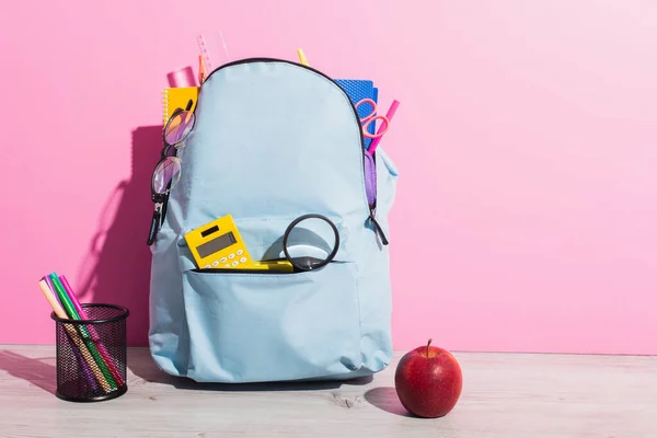 Sac à dos scolaire plein de papeterie près de pomme entière et porte-stylo avec stylos en feutre sur rose — Photo de stock