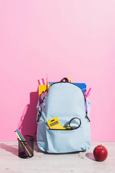 Blue backpack full of school supplies near ripe apple and pen holder with felt pens on pink — Stock Photo