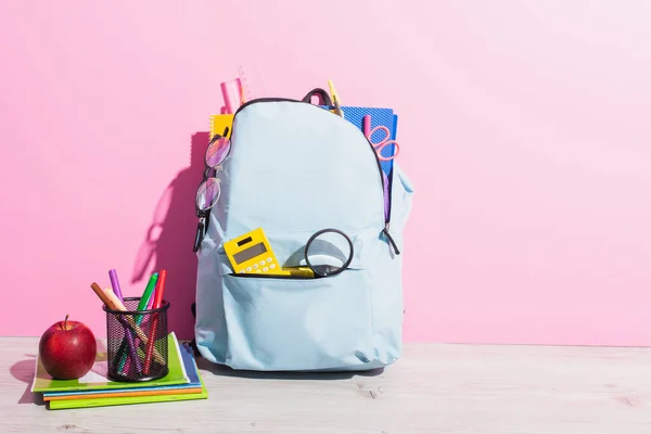 Zaino scuola pieno di cancelleria vicino a libri copia, mela e portapenne con pennarelli su rosa — Foto stock