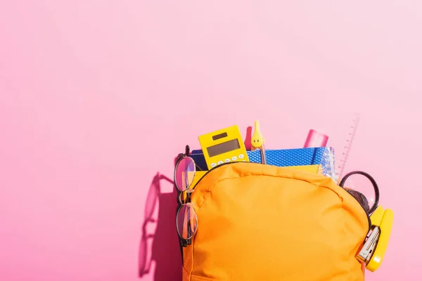 Zaino giallo pieno di cancelleria scolastica e occhiali su rosa — Foto stock