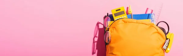 Horizontal image of yellow backpack packed with school supplies and eyeglasses on pink — Stock Photo