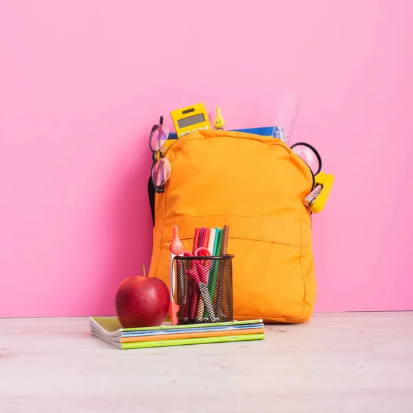 Mochila amarilla llena de útiles escolares cerca de cuadernos, porta bolígrafos con plumas de fieltro, tijeras y manzana madura en rosa - foto de stock