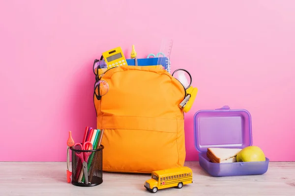 Zaino giallo con cancelleria scolastica vicino giocattolo scuolabus, portapenne e portapenne con pennarelli e forbici su rosa — Foto stock