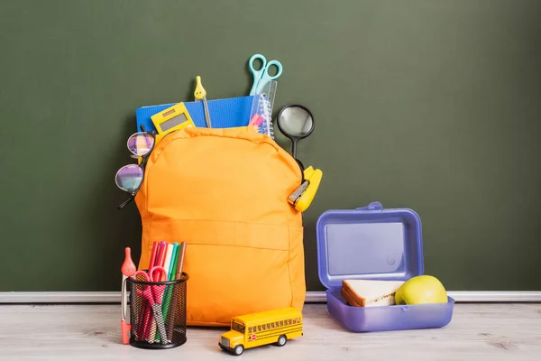 Gelber Rucksack voller Schulbedarf in der Nähe von Lunchbox, Schulbus-Modell und Stifthalter auf Schreibtisch neben grüner Tafel — Stockfoto