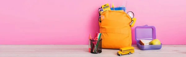 Horizontales Bild eines gelben Rucksacks mit Schulpapier in der Nähe von Lunchbox, Spielzeugschulbus und Stifthalter mit Filzstiften auf rosa — Stockfoto