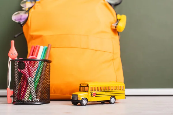 Sac à dos jaune près du modèle d'autobus scolaire, porte-stylo avec stylos en feutre, ciseaux et séparateur de boussole sur le bureau près du tableau vert — Photo de stock
