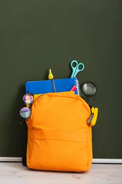 Mochila amarilla con útiles escolares en el escritorio cerca de pizarra verde - foto de stock