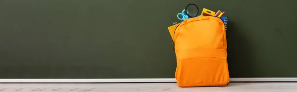 Imagem horizontal de mochila amarela cheia de artigos de papelaria escolar na mesa perto de quadro-negro verde — Stock Photo