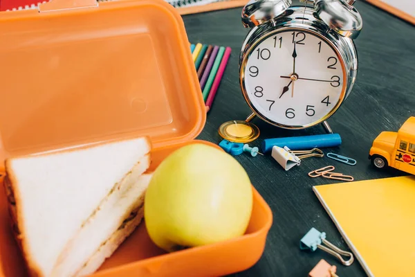 Focus selettivo del lunch box con gustosi panini e mela matura vicino sveglia vintage e forniture scolastiche su lavagna nera — Foto stock