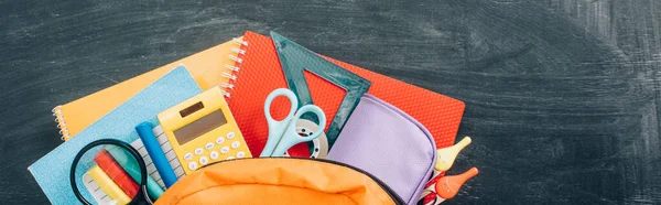 Top view of school backpack with stationery on black chalkboard, panoramic shot — Stock Photo