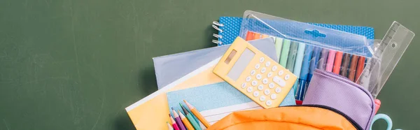 Top view of backpack full of school stationery on green chalkboard, horizontal image — Stock Photo